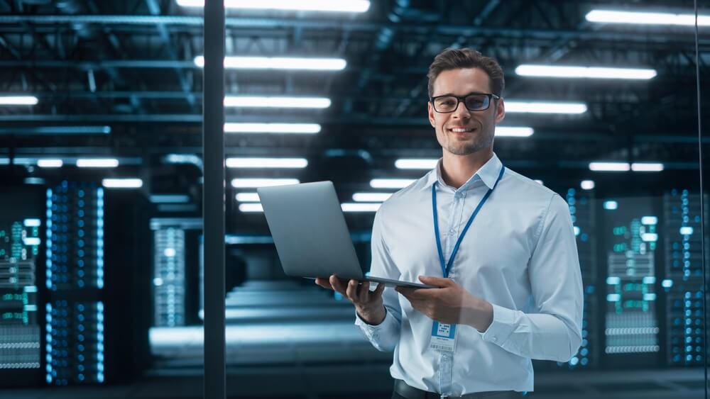 A man wearing glasses, a white shirt, and a blue lanyard stands inside a data center holding an open laptop. He smiles at the camera with server racks illuminated by lights in the background, showcasing the inclusive and diverse environment fostered by their commitment to DEI principles.