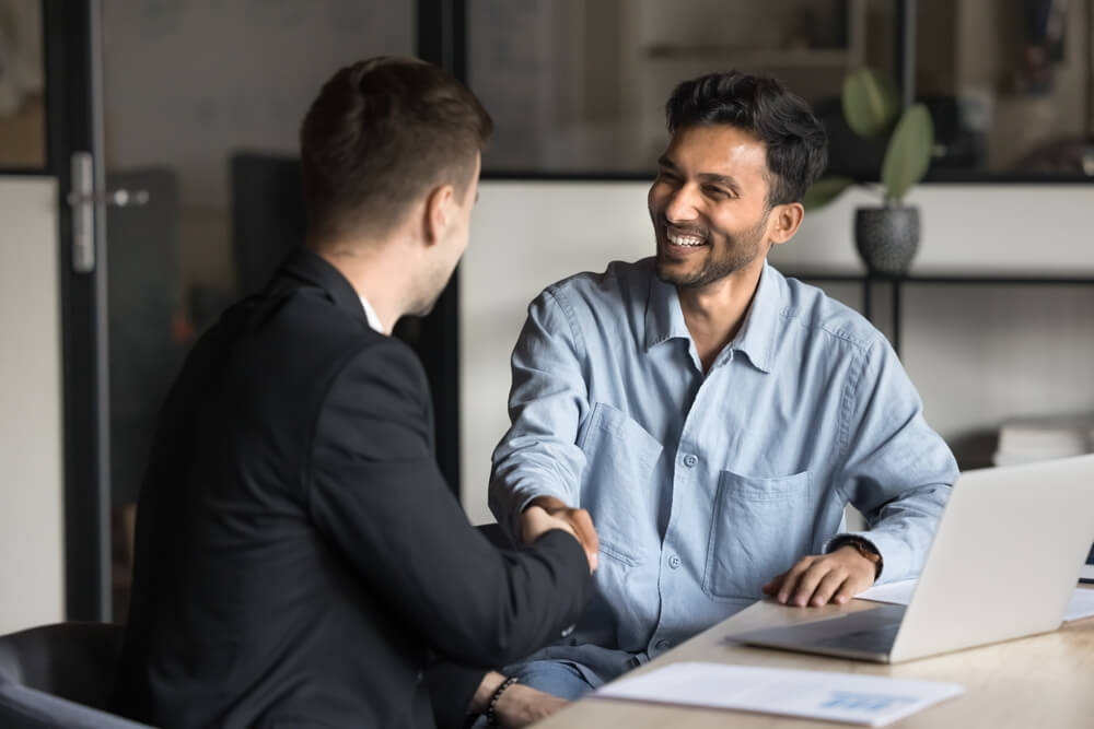 Two men in a professional setting are shaking hands and smiling. One man, dressed in a black suit, offers positive feedback to the other, who is wearing a light blue shirt. They are seated at a desk with a laptop, a document, and a potted plant in the background.