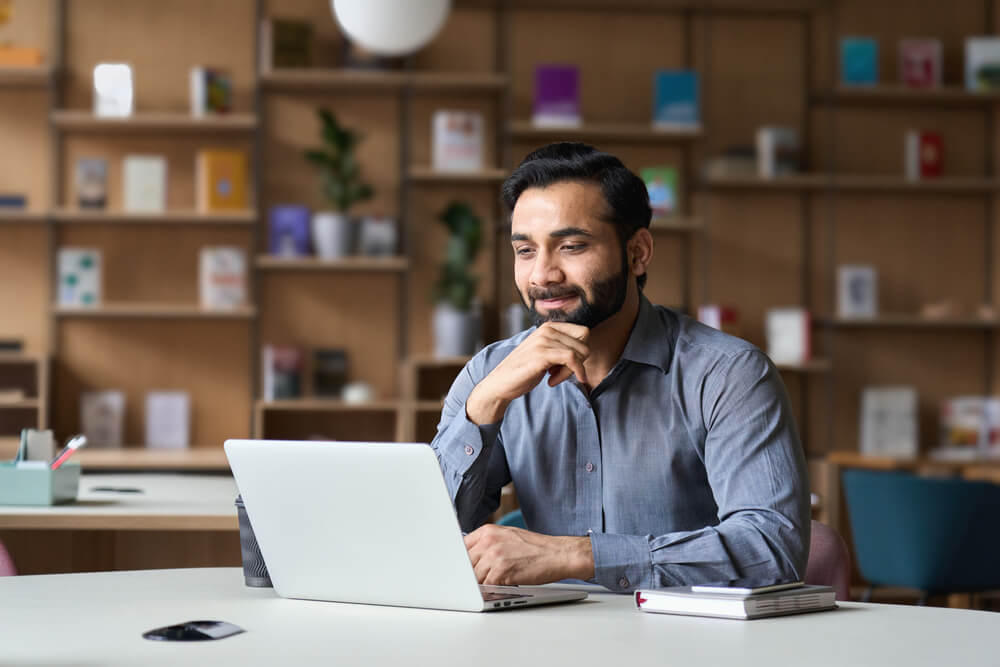 A man in a gray shirt sits at a table with a laptop, smiling and appearing focused. The background features bookshelves filled with government reports and various books, creating a cozy and studious atmosphere.