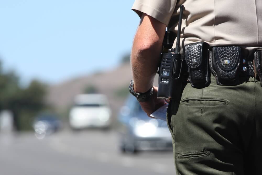 A police officer in uniform stands on the roadside, holding a notepad, with AI-integrated radio and belt accessories. Several cars are blurred in the background, highlighting the officer's cutting-edge tools.