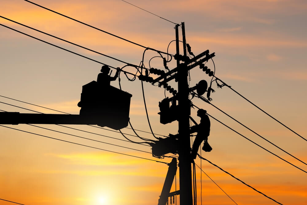 Two utility workers in silhouette on a power pole during a sunset, embodying timeless human effort even in an age shaped by AI. One is in a bucket truck, the other climbs the pole, both diligently working on electrical lines against a gradient sky of orange and yellow.