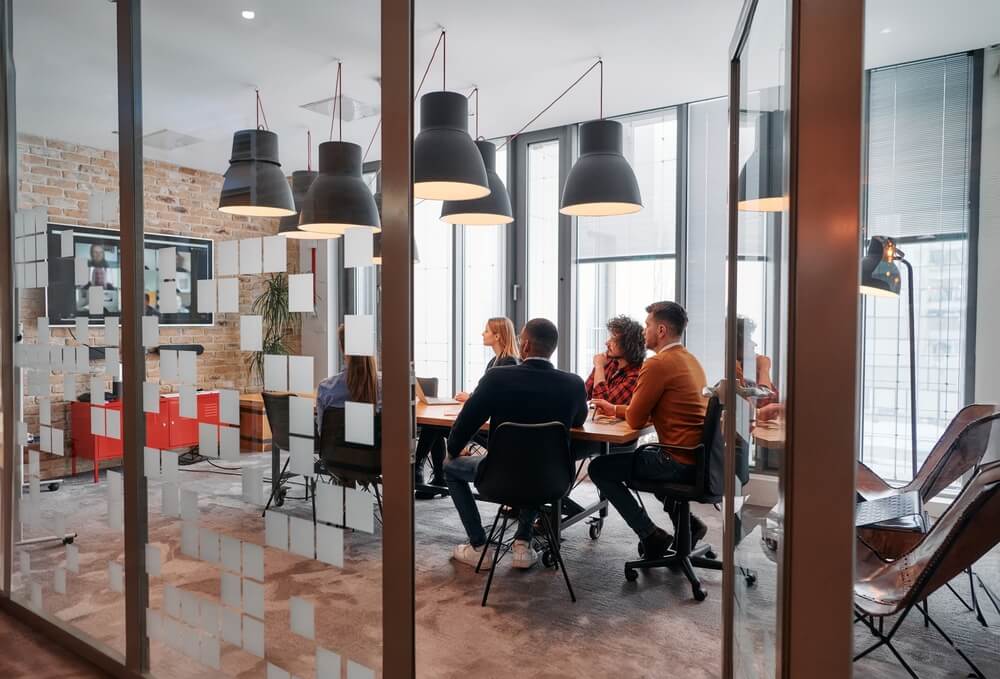 A diverse group of people sits around a conference table in a modern office with large glass walls, embodying the company's values. They engage in a meeting, focused on a screen on the wall. Hanging lights and contemporary decor create a professional atmosphere.