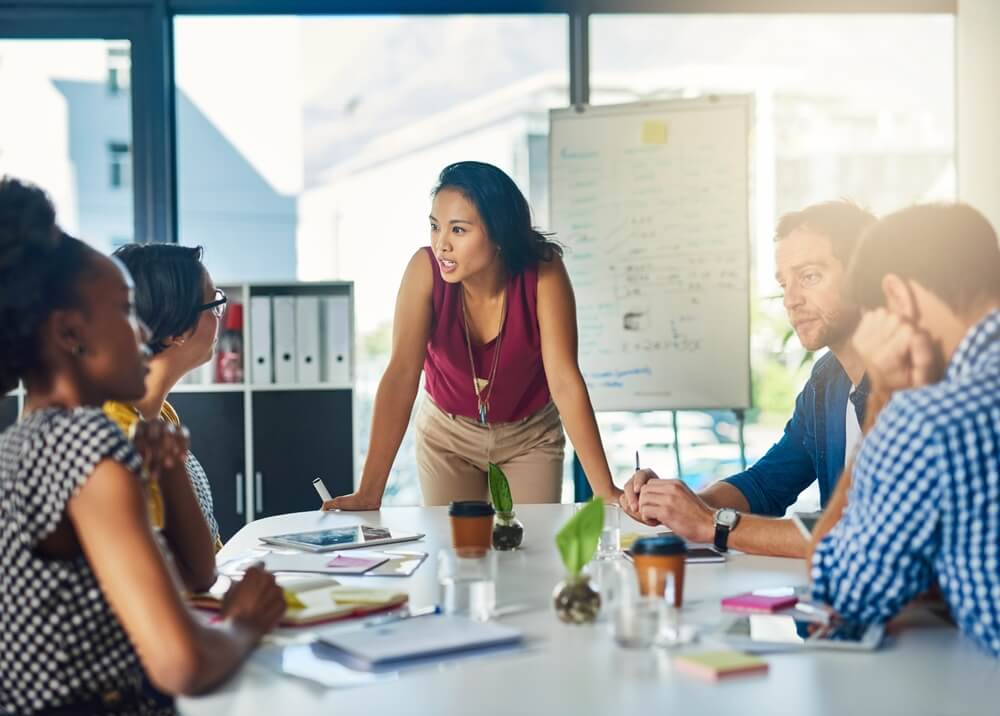 A woman in a red top stands at a conference table, illustrating the team's core values to engaged colleagues seated around. They are in a modern office with a whiteboard in the background. The table is cluttered with notes, coffee cups, and stationery.