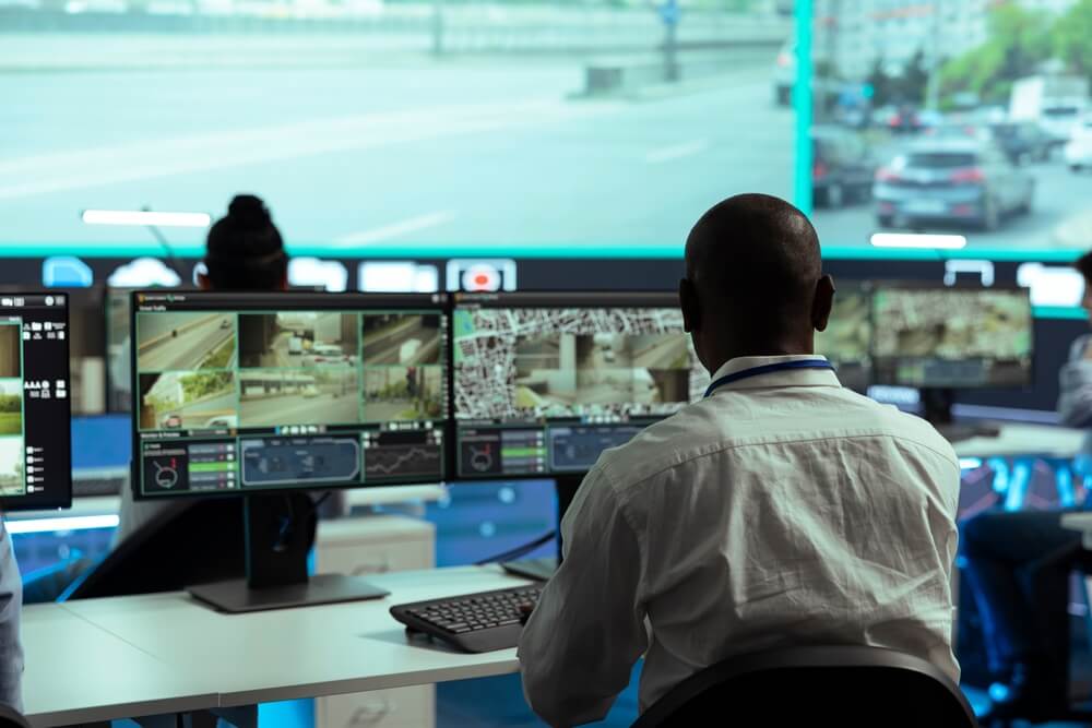 A person in a white shirt sits at a desk in front of multiple computer monitors displaying surveillance footage of diverse urban streets. The office environment is dimly lit, with a large screen dedicated to highlighting key DEI initiatives in the background.