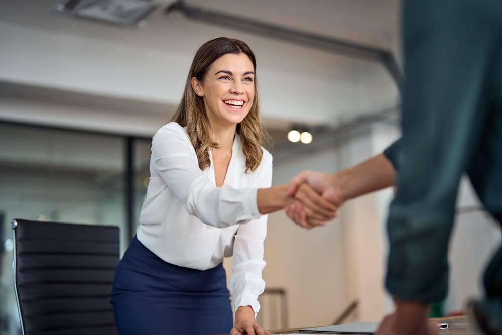 A woman in a white blouse and blue skirt smiles warmly, leaning forward to shake hands with someone across the table, embodying a culture of professionalism. She appears to be in an office setting, with a blurred background and a black chair nearby.