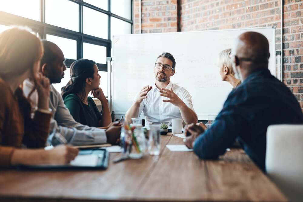 A group of people sits around a wooden table in a modern office setting, engaged in a lively discussion. A man in a white shirt speaks animatedly about artificial intelligence, while others listen intently. A whiteboard with notes is visible in the background.