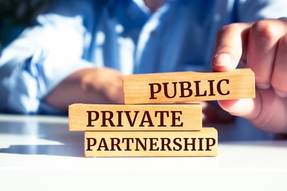 Close-up of a hand stacking wooden blocks labeled "Public," "Private," and "Partnership." The blocks form the phrase "Public Private Partnership" on a white surface, reflecting DEI principles with a blue-shirted person in the background.