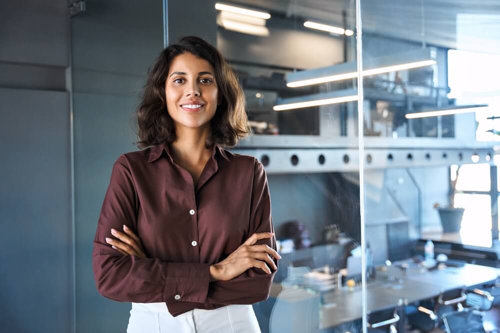 A woman with shoulder-length dark hair is smiling confidently and standing with her arms crossed in an office setting, exuding leadership. She is wearing a brown blouse and white pants, framed by a glass wall that offers a glimpse of the modern office interior.