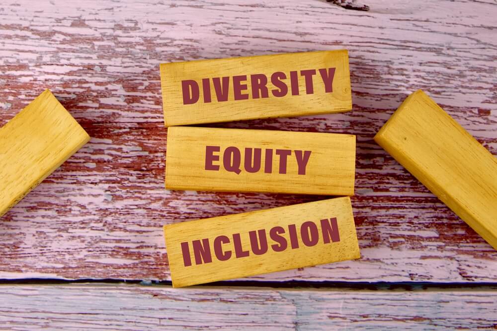 Wooden blocks on a painted wooden surface boldly showcase the words "Diversity," "Equity," and "Inclusion" in striking red letters, encapsulating the essence of DEI.