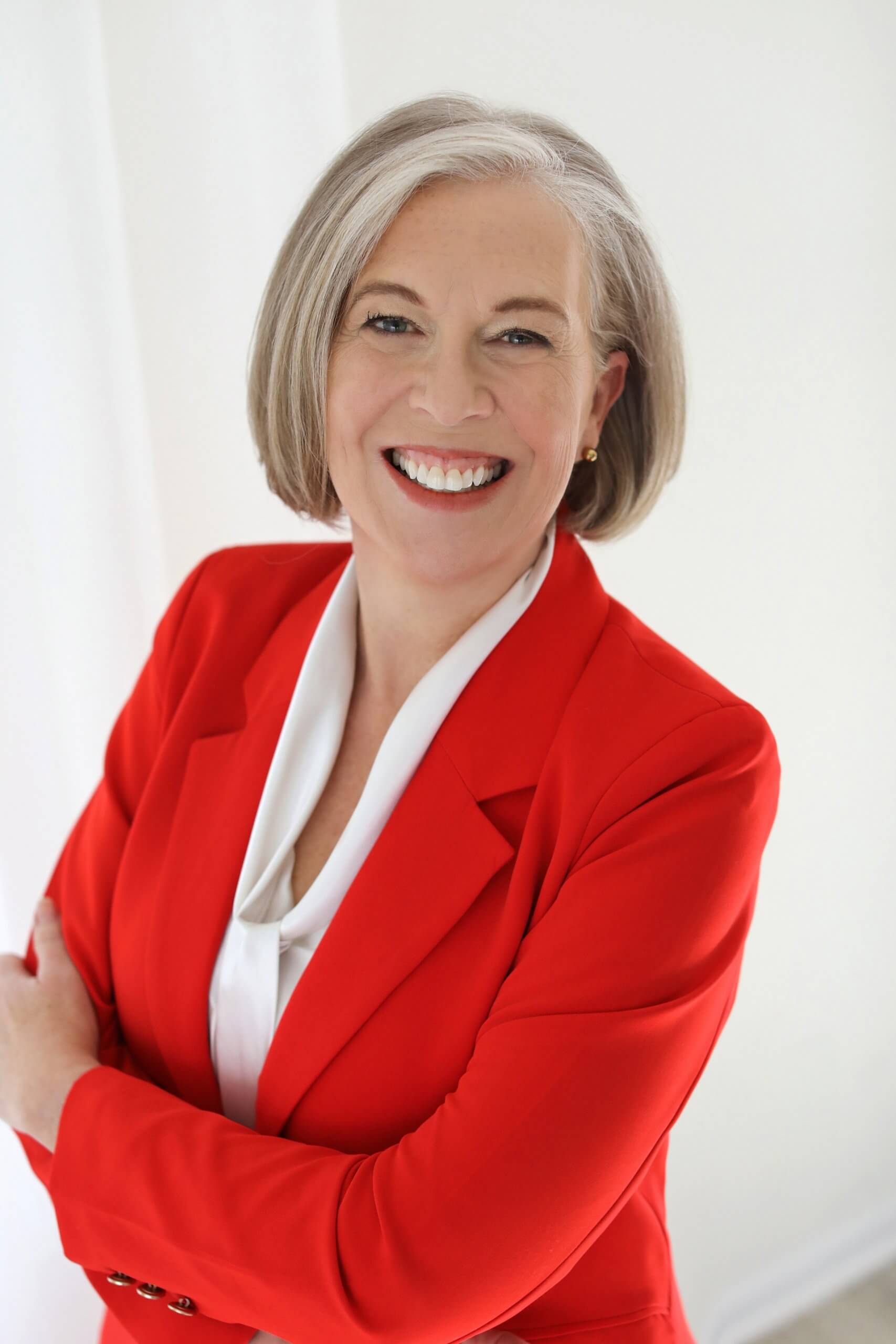 A smiling woman with short gray hair is wearing a red blazer over a white blouse. She is standing against a light background with her arms crossed.