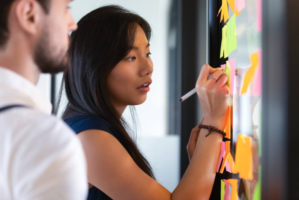 Two people stand in front of a board filled with colorful sticky notes. One writes on a note with a marker, while the other observes. Their focused collaboration suggests a brainstorming session, possibly enhanced by insights from artificial intelligence.