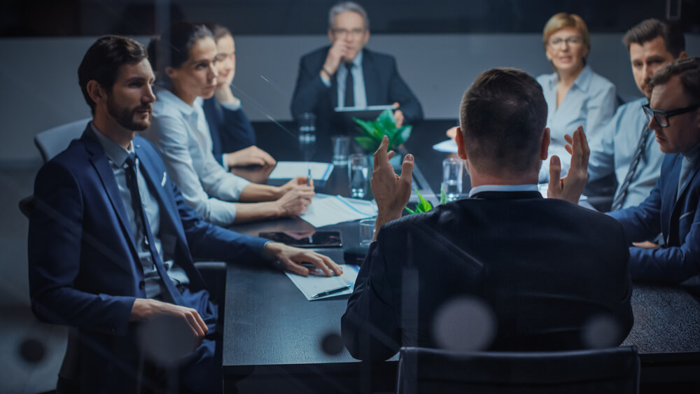 A group of business professionals sits around a conference table in a modern office, engaged in an important meeting. Some are diligently taking notes while others listen attentively, surrounded by glass partitions and lush potted plants that add vibrancy to the productive atmosphere.