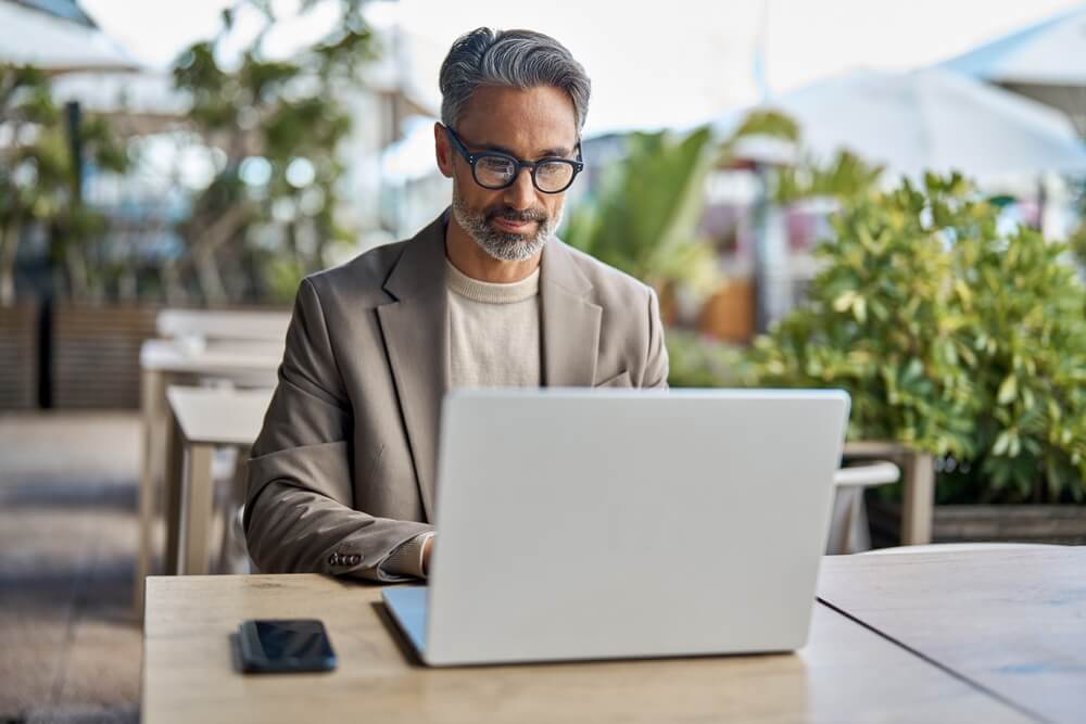 A man with glasses and gray hair, exuding leadership in his beige blazer, sits at an outdoor wooden table, focused on his laptop. A smartphone lies beside him, while lush greenery frames the scene.