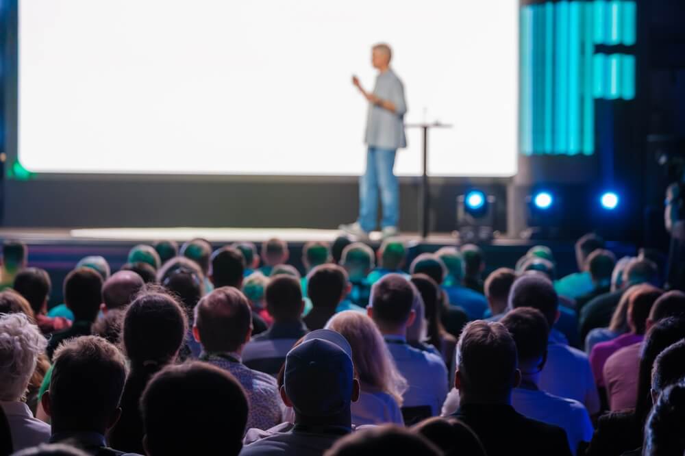 A leader stands on stage addressing an audience in a dimly lit auditorium. The attentive crowd is seated, and the brightly illuminated stage highlights the speaker against a large, mostly white backdrop, as blue and green lights add ambiance.