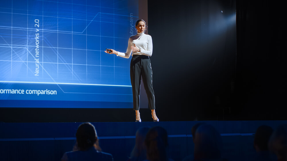 A person stands on stage giving a business presentation, holding a pointer while facing an attentive audience. Behind them, a large screen displays graphs and technical text related to neural networks.