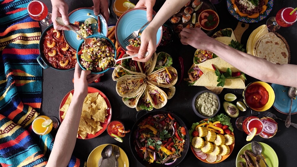 A colorful spread of Mexican dishes on a dark table, featuring tacos, nachos, grilled vegetables, dips, and assorted drinks. Multiple hands are reaching in to serve themselves while an AI-assisted playlist sets the energetic mood. A vibrant striped cloth decorates the side.