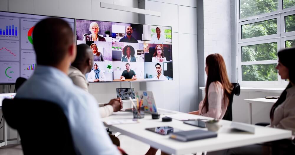 People in a modern conference room engage in a video call displayed on large screens. The space, marked by leadership and innovation, features large windows, white tables, and digital charts on screens. Laptops and notepads are on the table, indicating a dynamic business meeting.