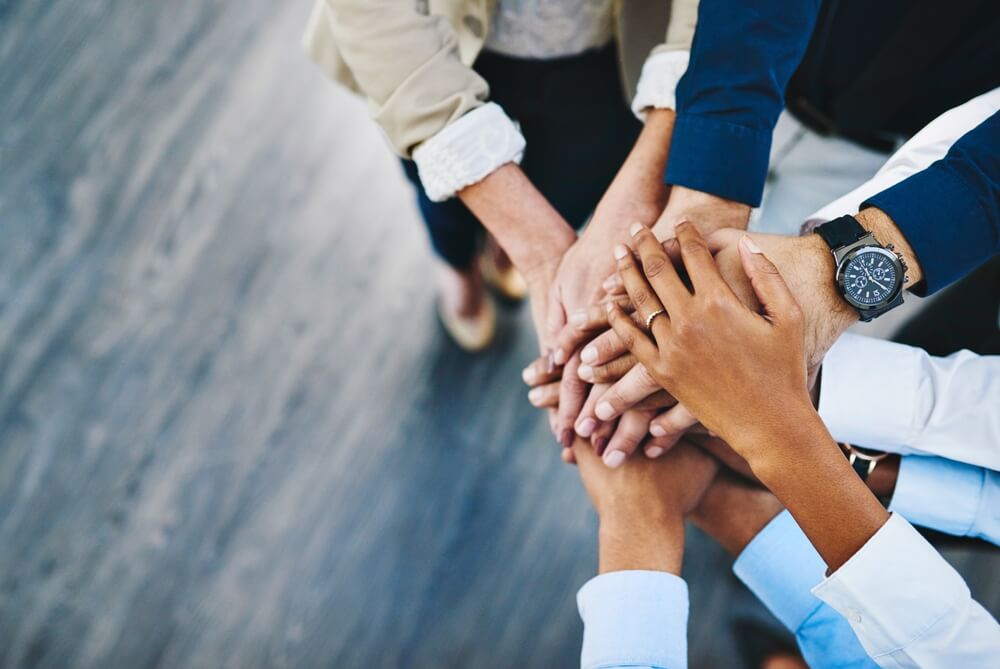 A diverse group of people, including government officials, stands in a circle with their hands stacked together in the center, symbolizing teamwork and unity. The background is a wooden floor, and individuals are wearing a blend of casual and business attire.