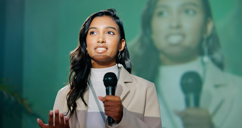 A woman, exuding the poise of a leader, holds a microphone and speaks on stage with her image projected in the background. Wearing a light blazer, she addresses an audience confidently against a green backdrop.