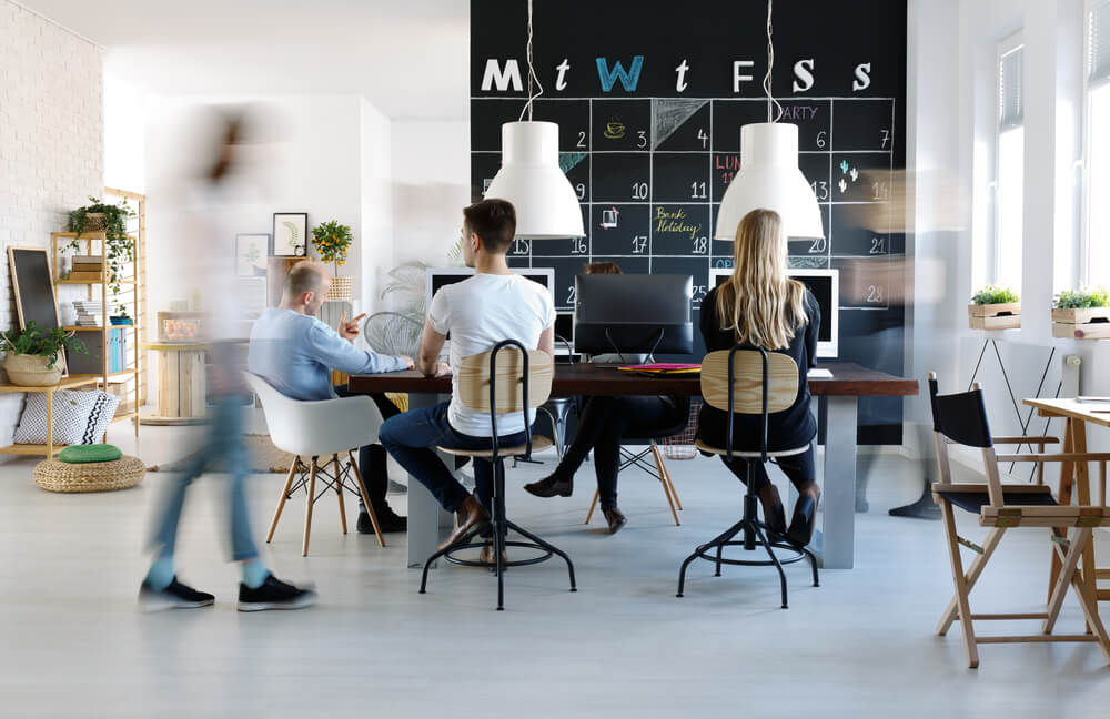 A modern office space buzzes with vision as people work and walk around. A blackboard wall displays a calendar, while computers, chairs, and plants on tables foster a dynamic and collaborative environment.