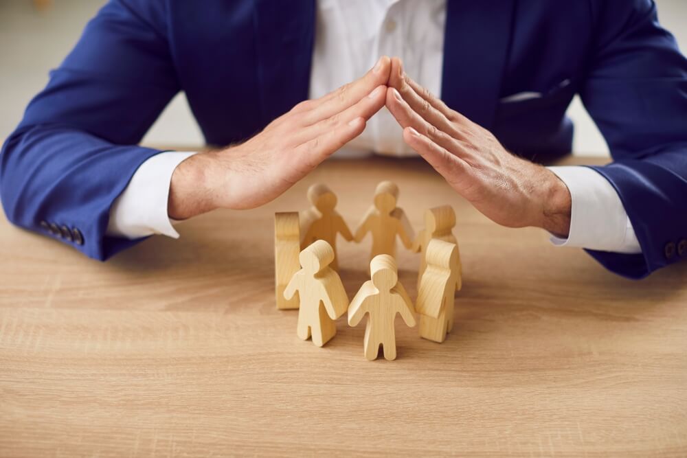 A person in a suit sits at a table, hands forming a protective dome over wooden cutouts of people, symbolizing management's role in safeguarding and caring for a community.