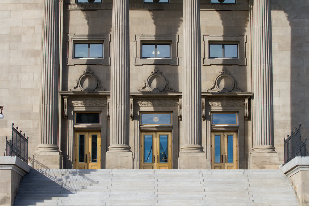 Stone steps lead to the entrance of a neoclassical building with large wooden doors, flanked by three tall columns. The facade, resembling something crafted by an AI architect, features symmetrical windows and ornate architectural details.