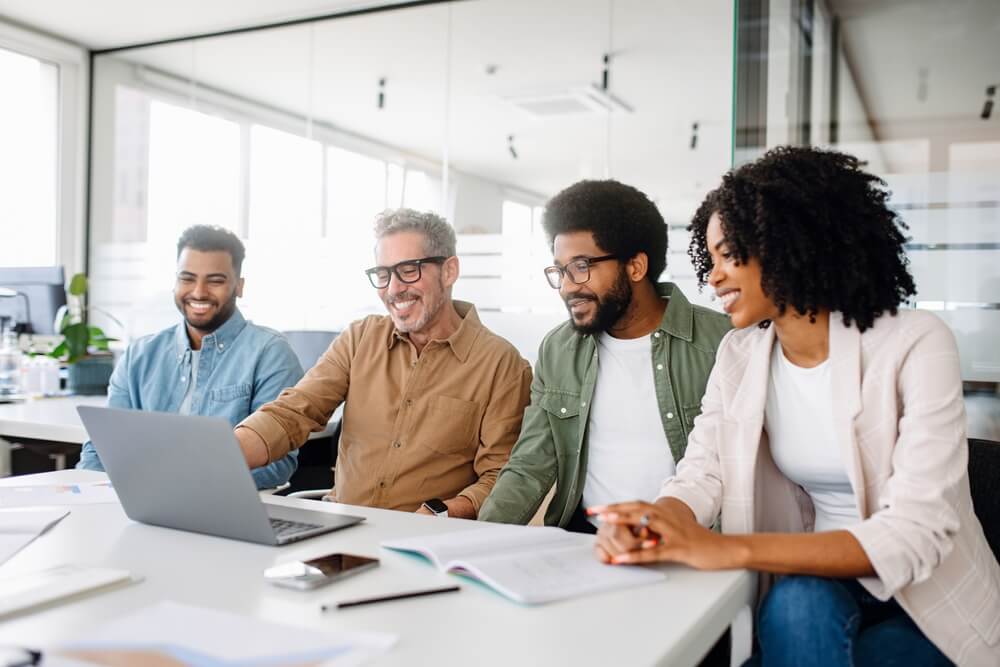 A diverse group of four people sit around a table with a laptop, smiling and engaged in a collaborative work environment. The modern office, with its large windows, sets the scene for leadership to flourish as books and documents spread across the table inspire innovative ideas.