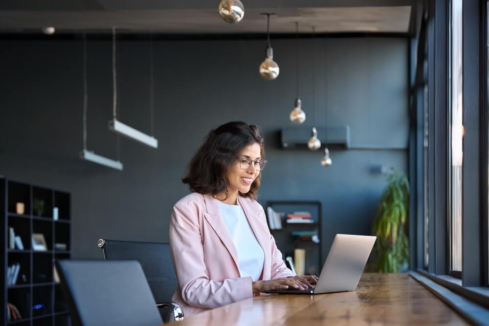 A woman in a pink blazer sits at a long wooden table, working on her laptop in a bright, modern workplace. She appears focused and happy amidst large windows, pendant lights, and a well-stocked bookshelf in the background.