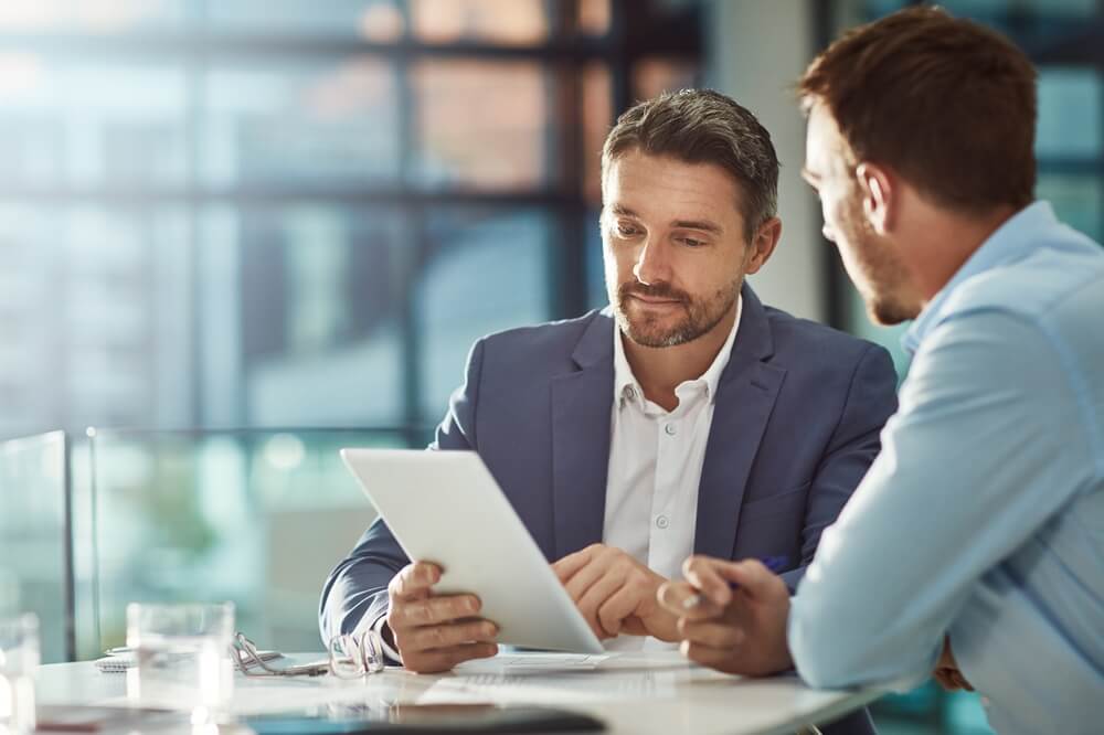 Two men sitting at a table in a modern office workplace are engaged in a discussion. One is holding a tablet, and they both appear focused on its screen. Sunlight streams through large windows in the background, creating a bright, professional atmosphere.