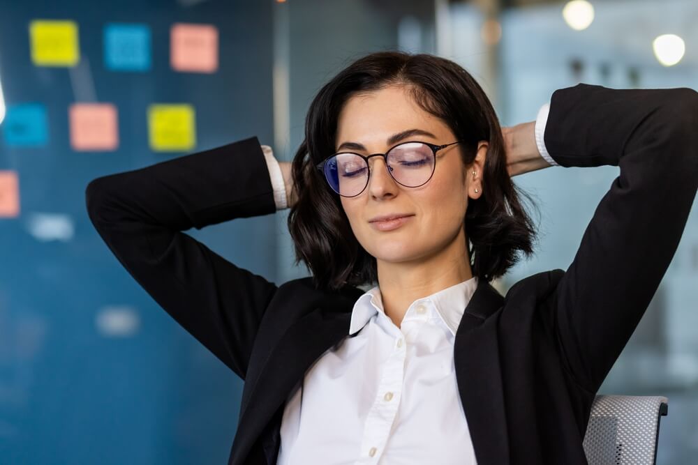 A woman in a business suit, embodying leadership, leans back in her chair with hands behind her head and eyes closed, exuding relaxation. In the background, a glass wall adorned with colorful sticky notes is visible.