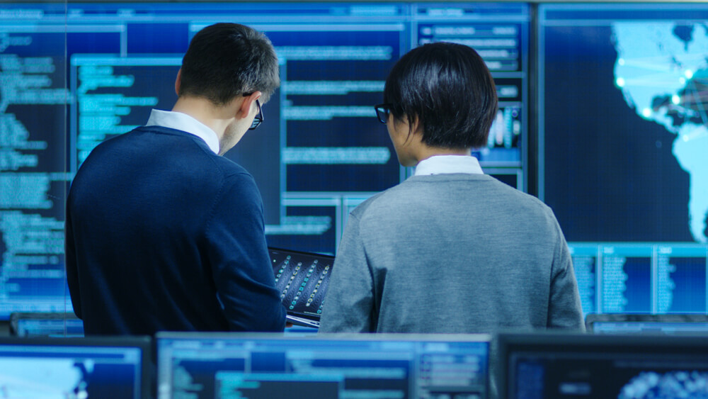 In a tech environment, two individuals analyze data on multiple screens, with one holding a tablet. The background's digital maps and projections hint at government cybersecurity or data analysis work. Both are wearing glasses, focused and intent on their task.