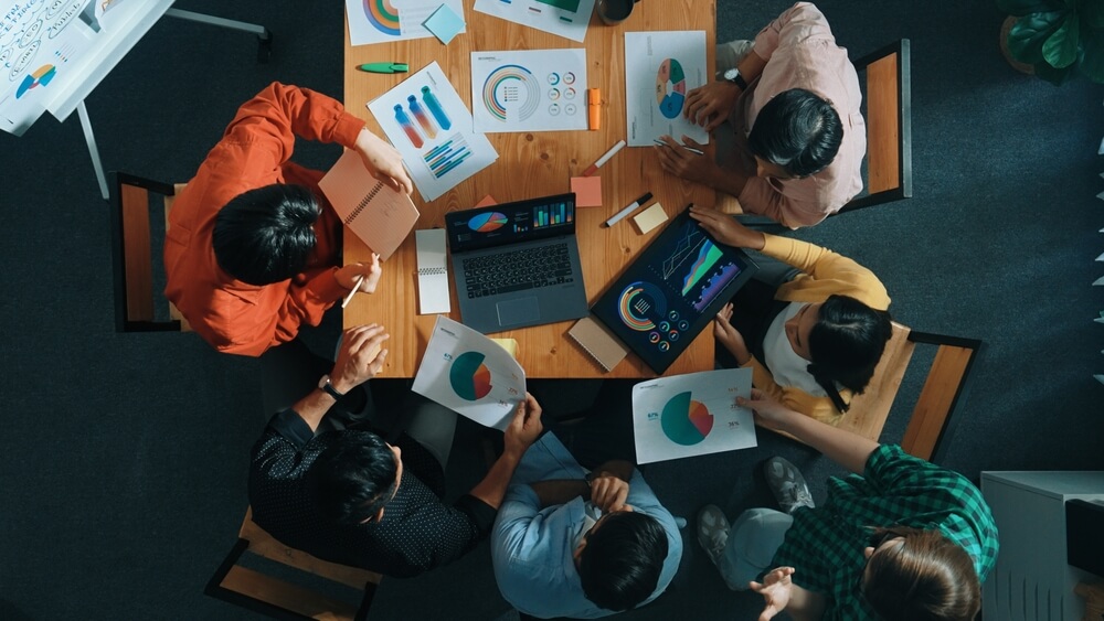 Aerial view of seven people gathered around a table, discussing colorful charts and graphs that highlight well-being metrics. A laptop and papers with pie and bar charts are visible, suggesting a collaborative business meeting focused on holistic team discussions.