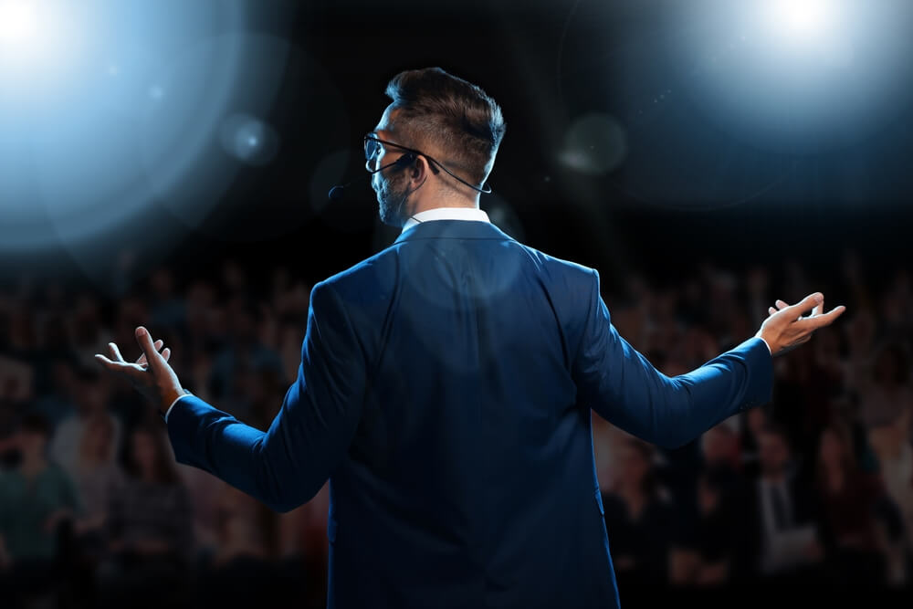 A man in a blue suit and glasses speaks into a headset microphone on stage, passionately gesturing with both arms. He's addressing the blurred audience under bright stage lights, creating lens flares, as he discusses the importance of well-being.